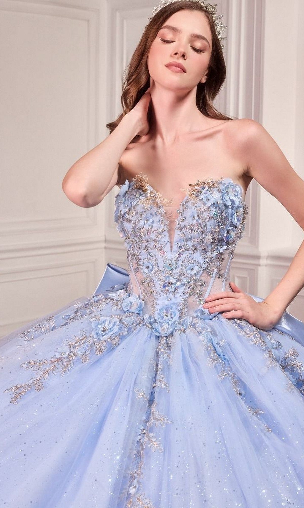 A woman models the Quinceanera Dress 15726 by Ladivine, a strapless light blue ball gown with 3D flowers and gold accents, posing indoors against a white paneled wall.